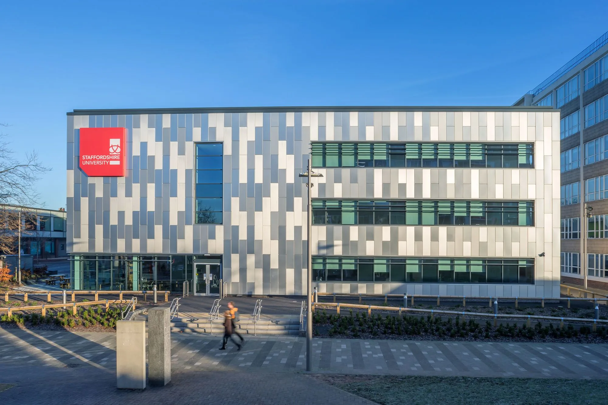 Exterior view of a modern Staffordshire University building featuring a geometric grey and white patterned facade, large horizontal windows, and a red logo sign.