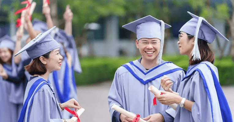 Three smiling university graduates in grey and blue gowns and mortarboards hold diplomas during an outdoor graduation celebration.