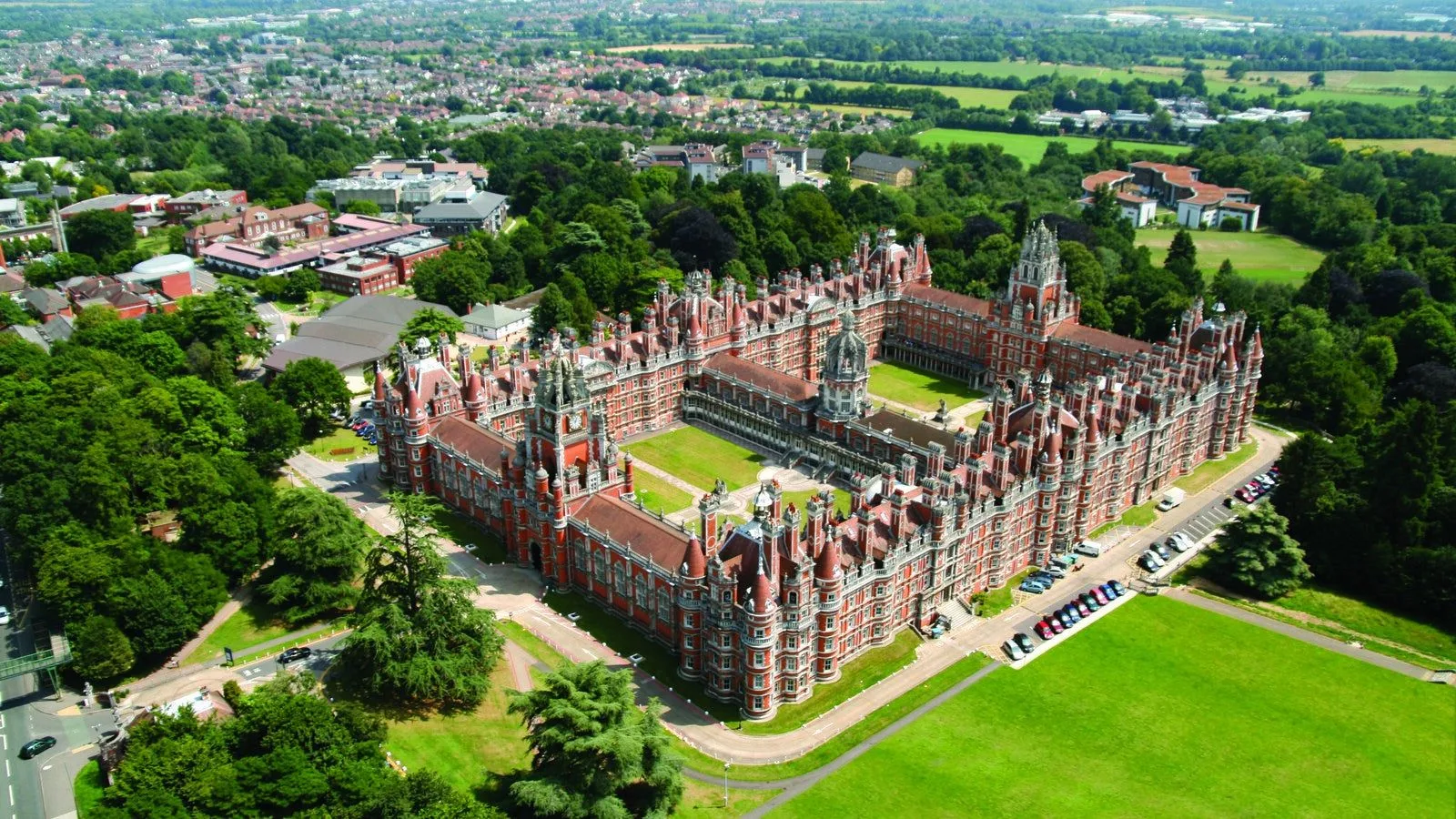 Aerial view of Royal Holloway, University of London’s historic red-brick Founder’s Building, surrounded by green lawns and trees.
