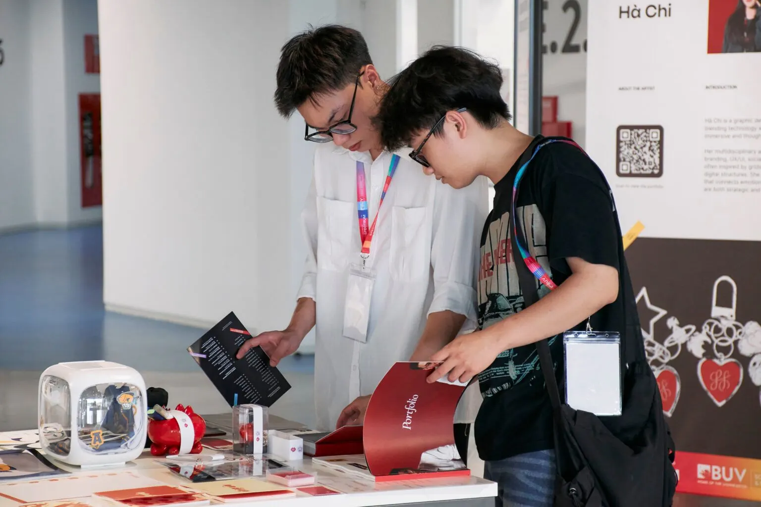 Two young men examine materials at a university event display table, with brochures, small objects, and BUV promotional signage visible in the background.