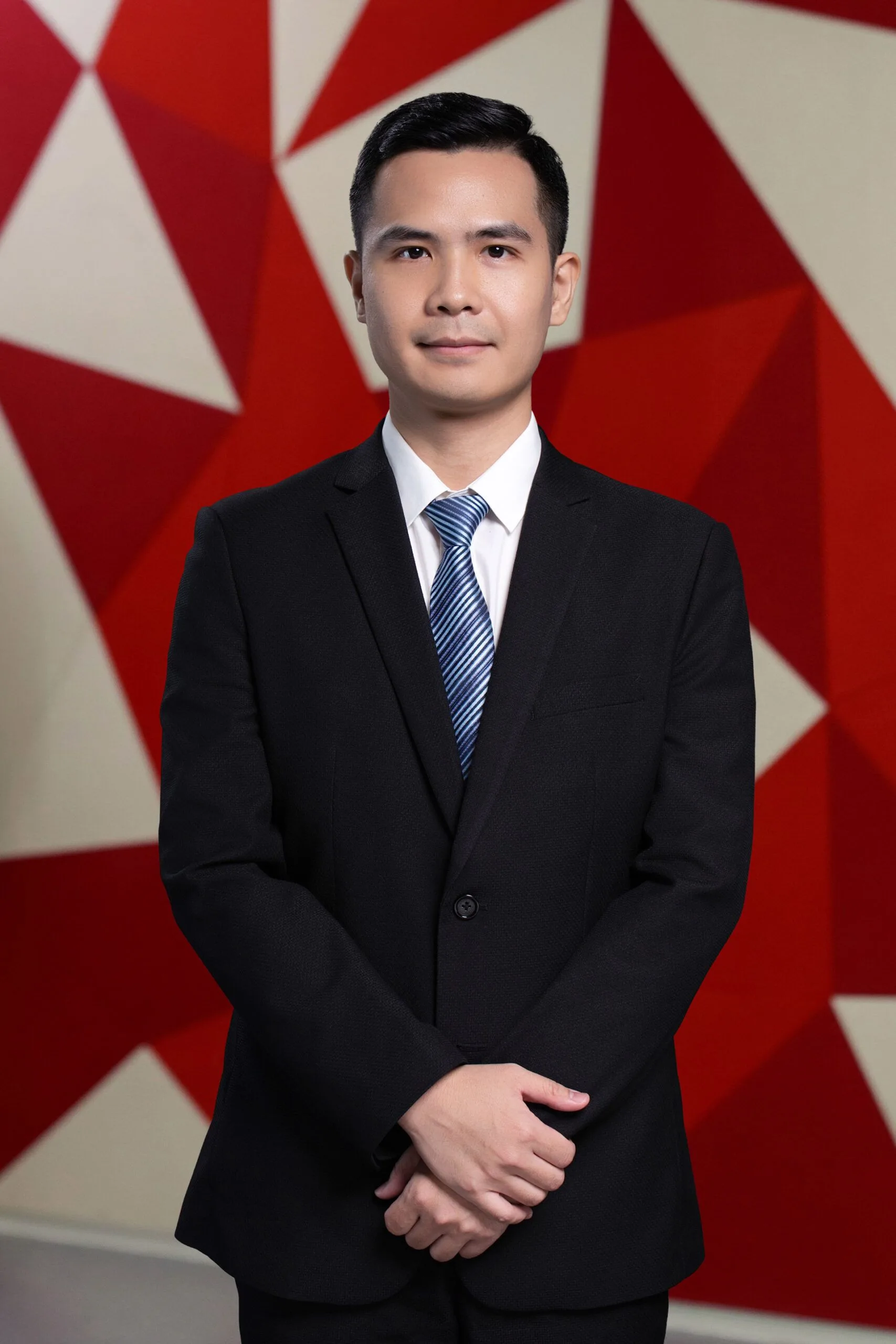 Portrait of Dong Manh Cuong wearing a black suit and blue striped tie, standing with his hands clasped against a red-and-white geometric backdrop.