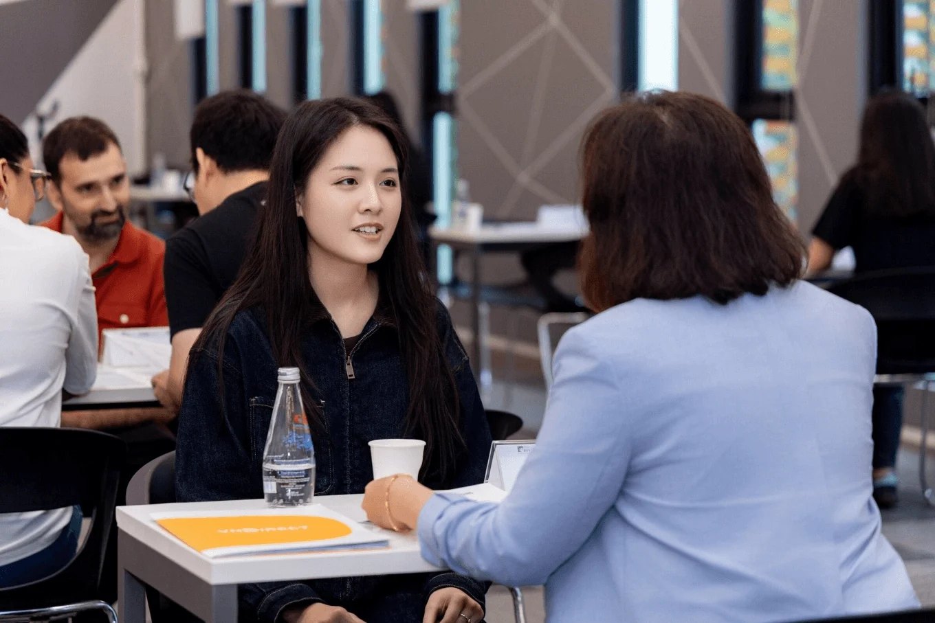 A young woman in a university setting talks with someone at a table during a discussion, with notebooks and drinks in front of them.