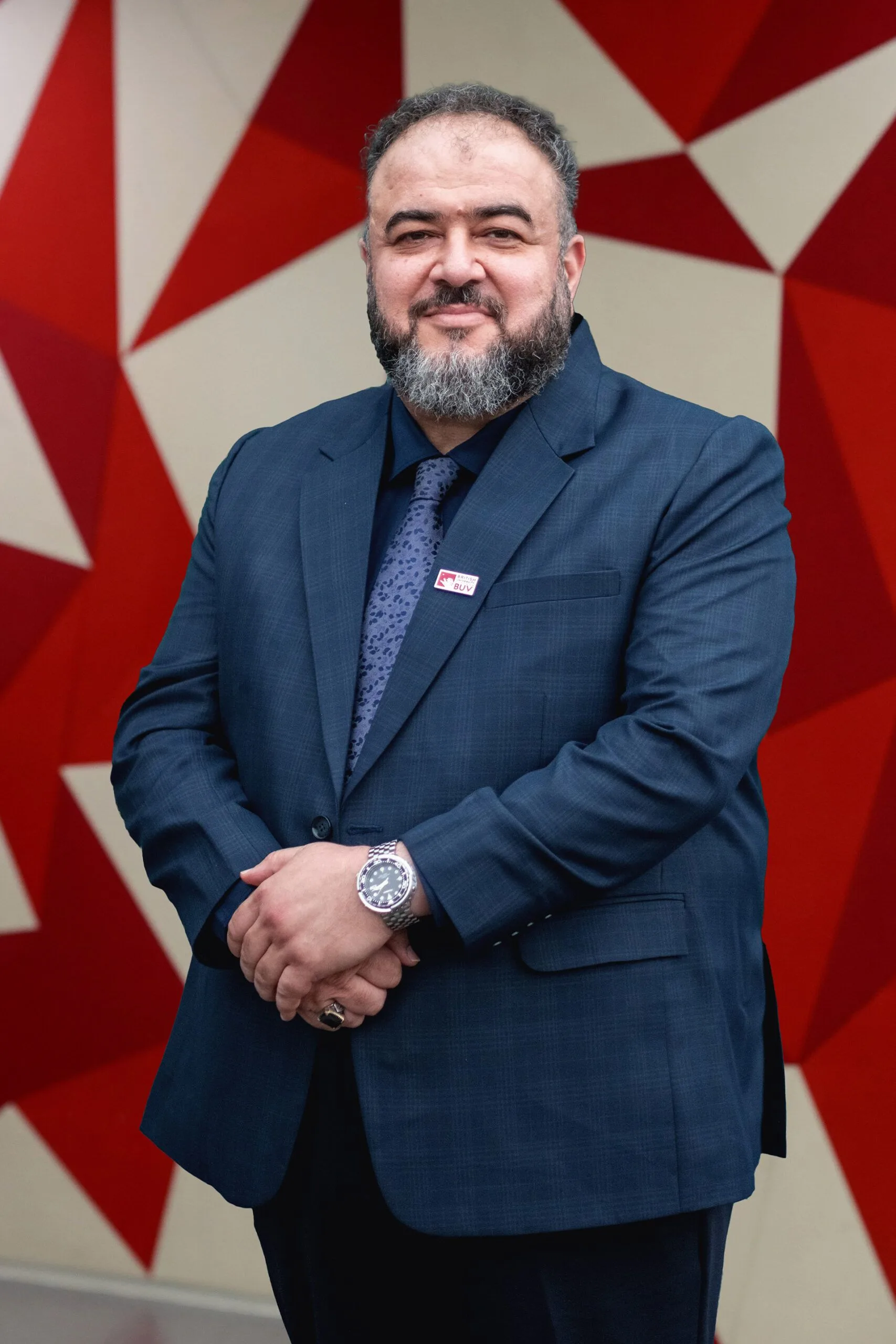 Portrait of Ali A. Dulaimi standing and smiling in a navy suit against a red geometric backdrop.