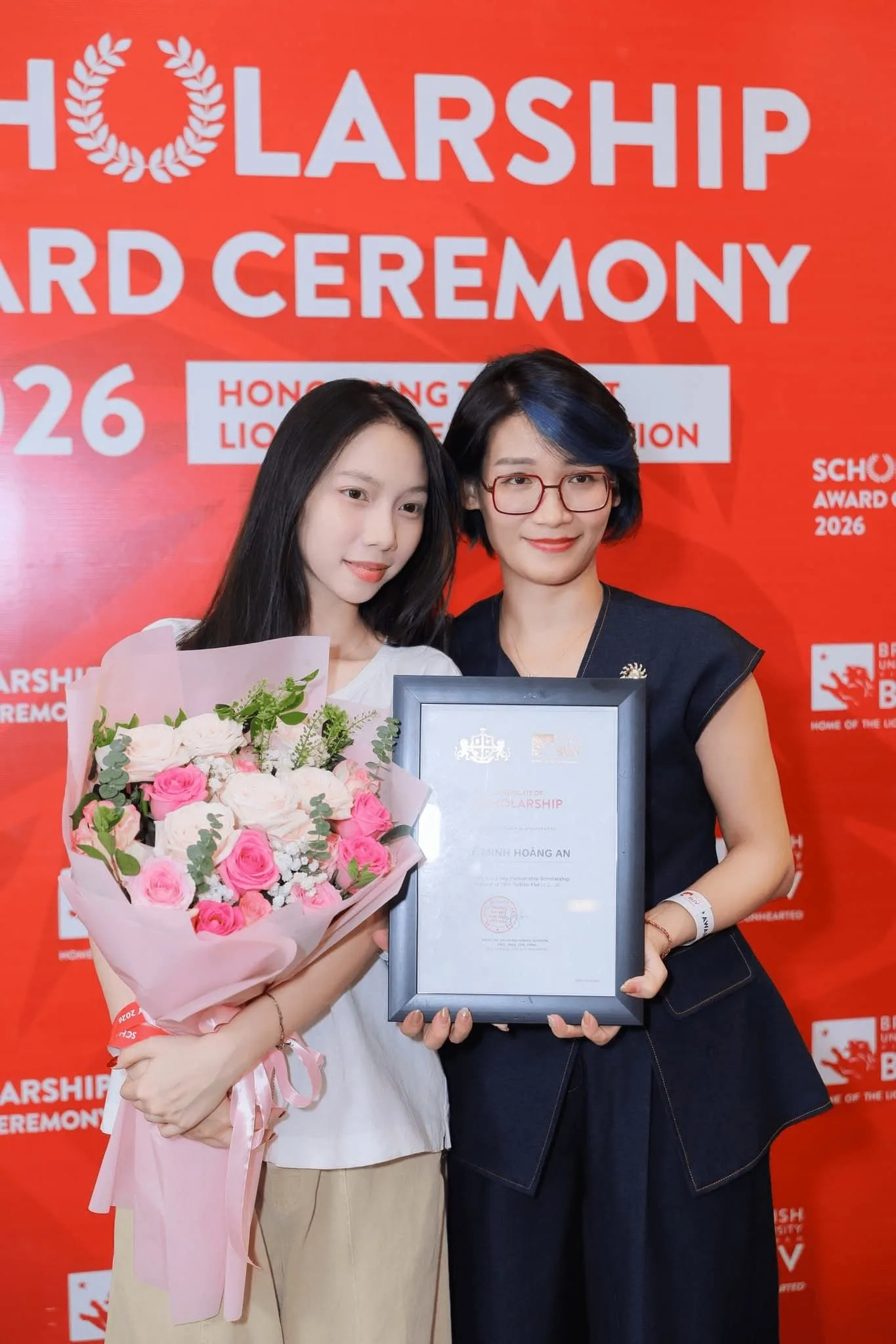 Two women pose at a 2026 scholarship award ceremony; one holds a bouquet of pink and white flowers while the other displays a framed certificate against a red event backdrop.