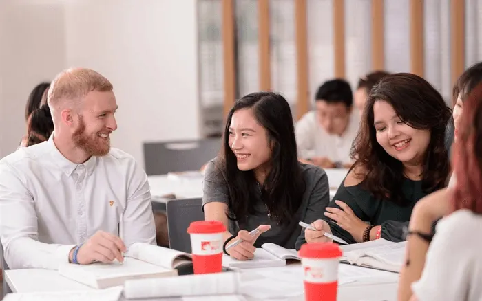A group of international students sit together at a table in a classroom setting, smiling and chatting while studying.