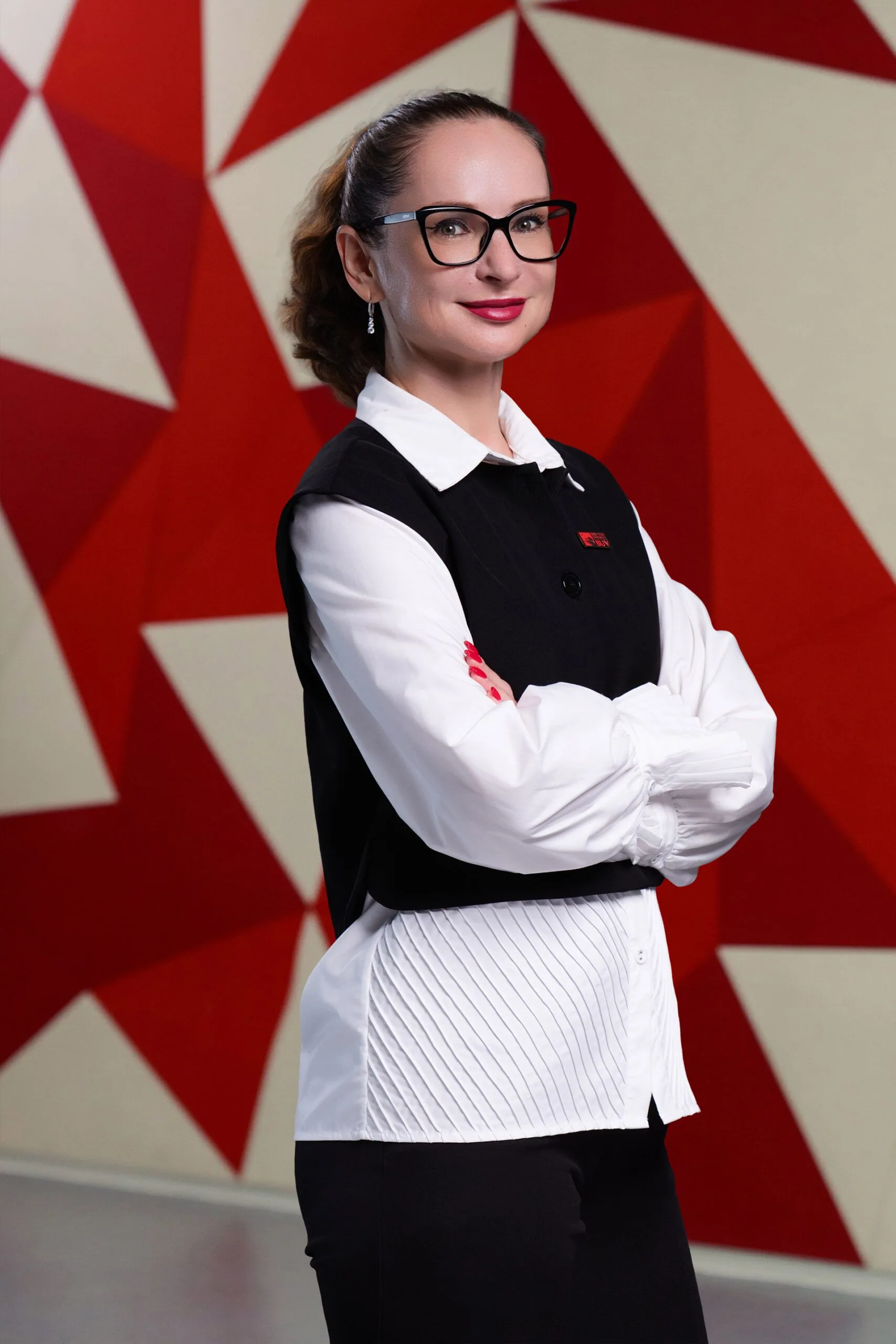 Portrait of a woman (Yulia) standing with arms crossed in front of a red and white geometric backdrop, wearing a black vest over a white shirt and glasses.