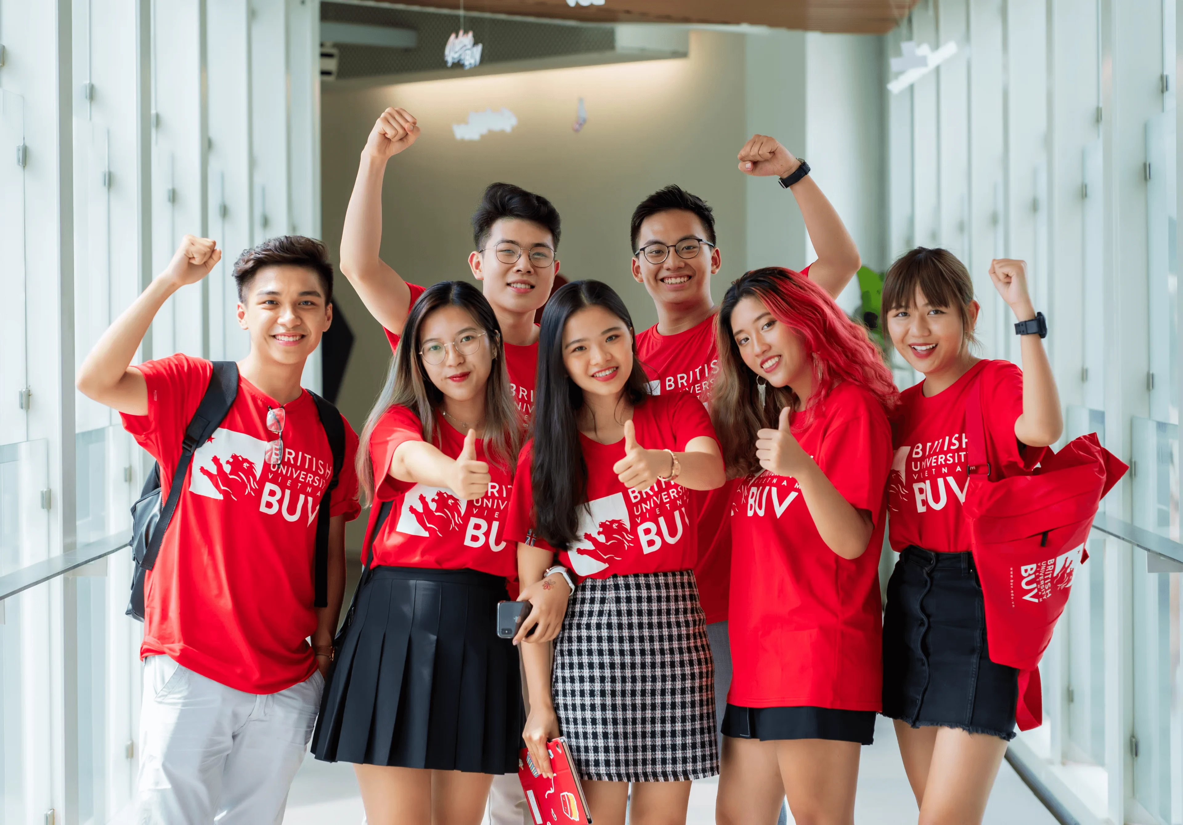 Seven BUV students in red university T-shirts smile, raise their fists, and give thumbs-up in a bright campus hallway.