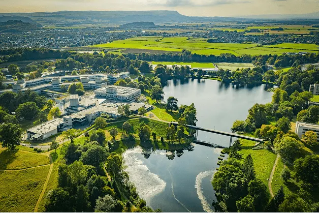Aerial view of the University of Stirling campus, surrounded by a lake, lush greenery, and countryside.