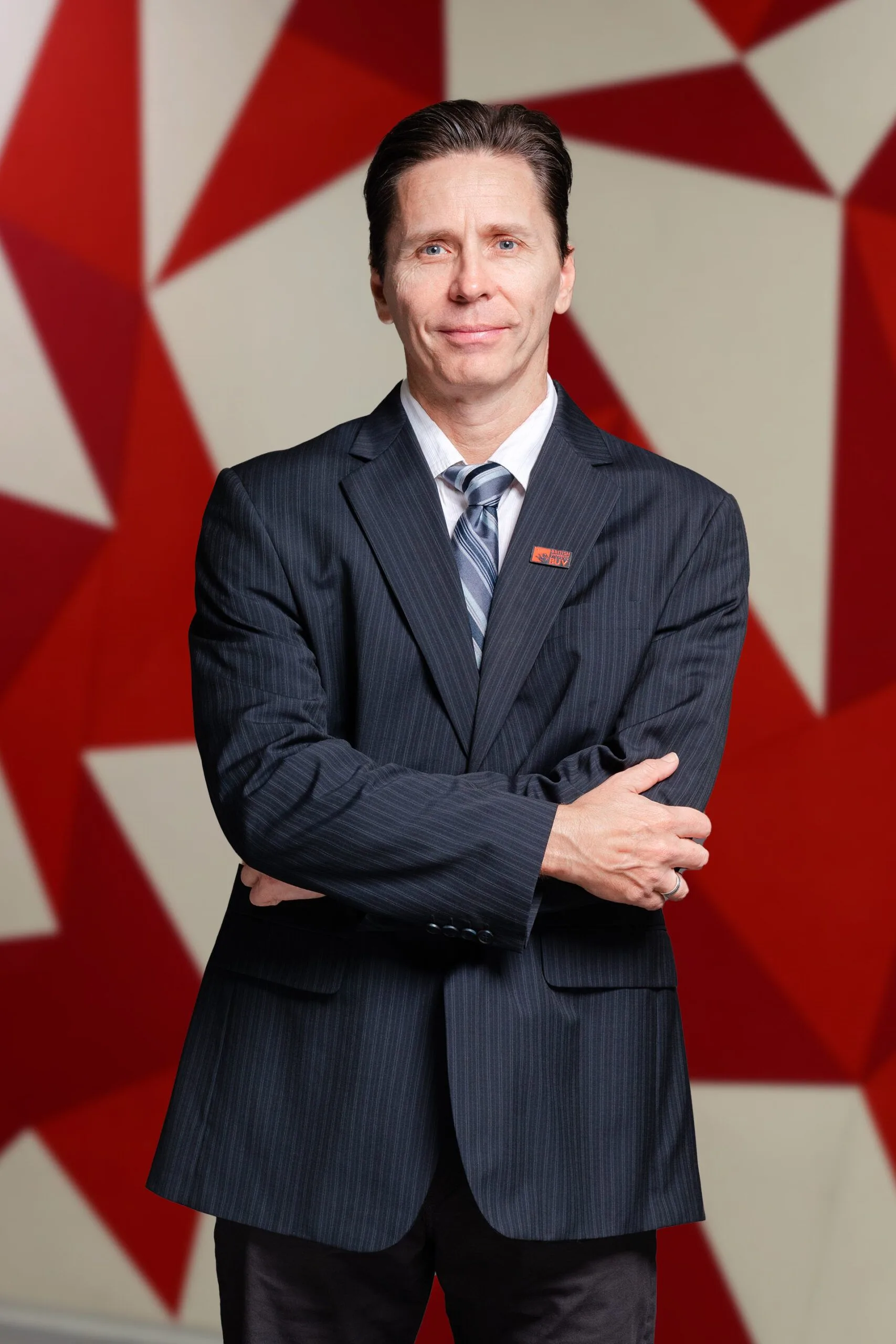 Portrait of Ross Adrian (DR-Ross-Adrian), standing with arms crossed in front of a red and white star-pattern backdrop.