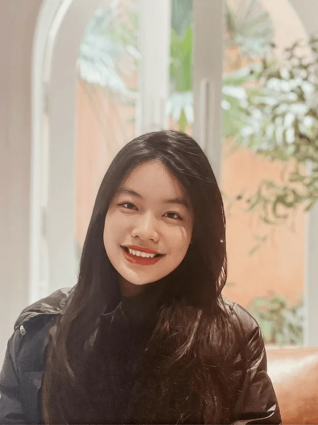 Portrait of Ha Vy smiling indoors, wearing a black turtleneck and jacket, with long dark hair and a softly lit arched window in the background.