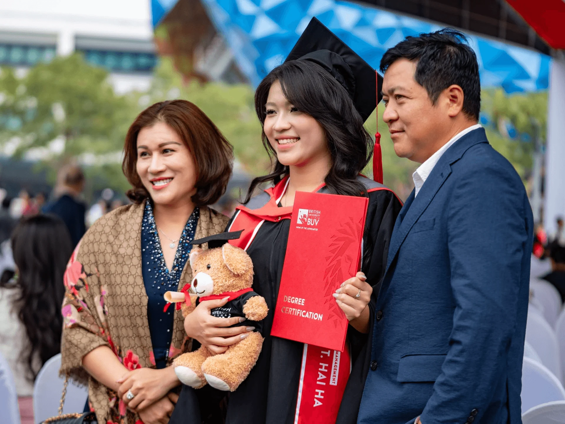 BUV graduate in cap and gown celebrates graduation with parents, holding a red degree certification folder