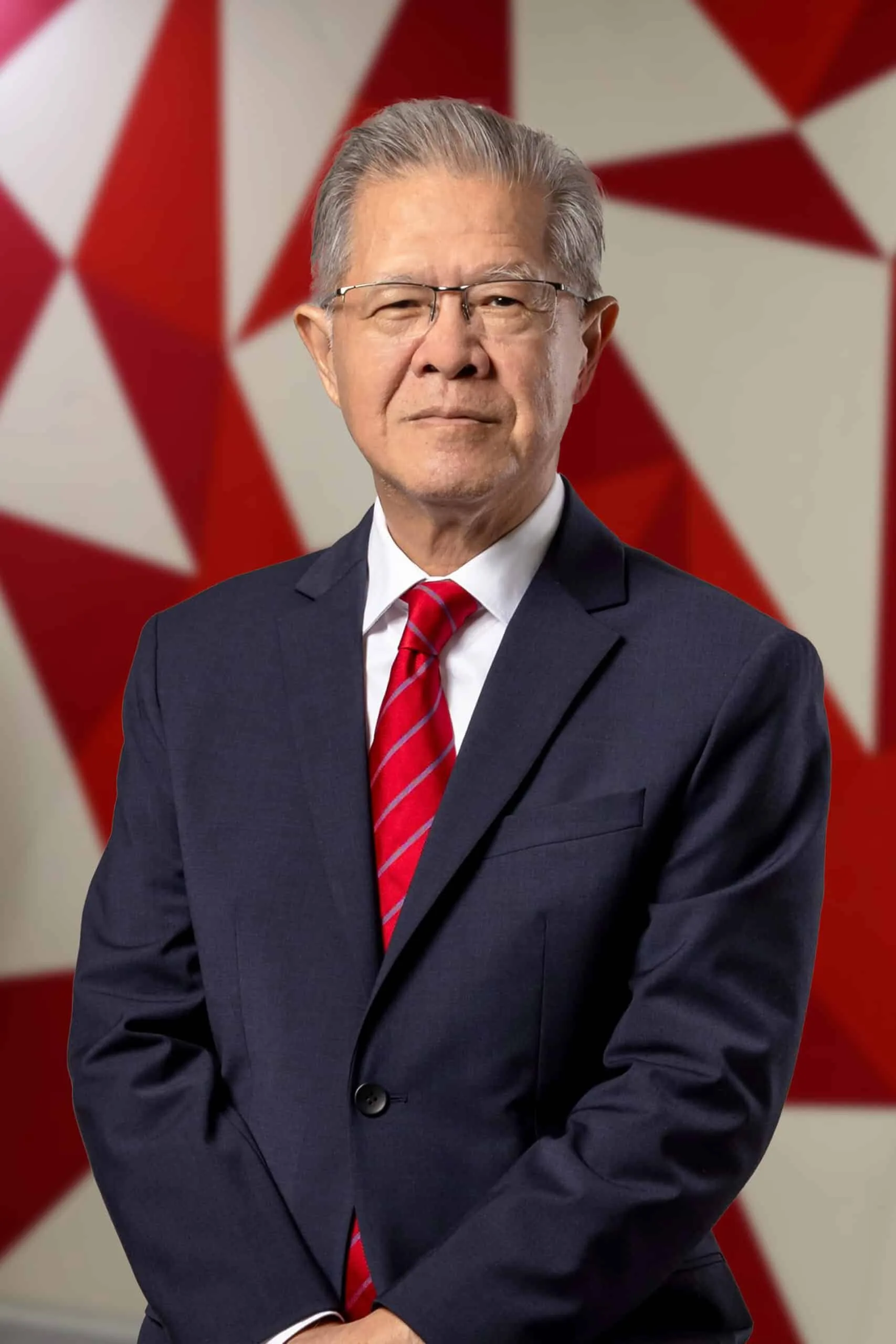 Portrait of Teo Tong Khooi in a dark suit and red-striped tie, standing in front of a red-and-white geometric backdrop.