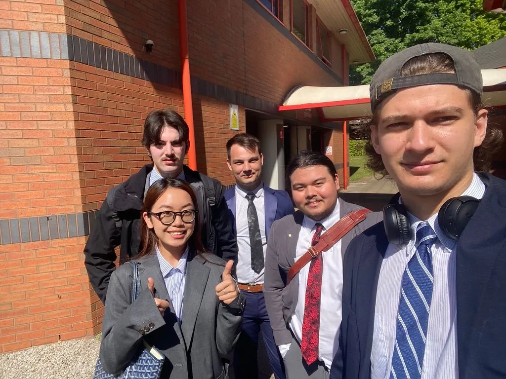 Five young adults dressed in formal attire pose for a sunny group selfie outside a red brick building