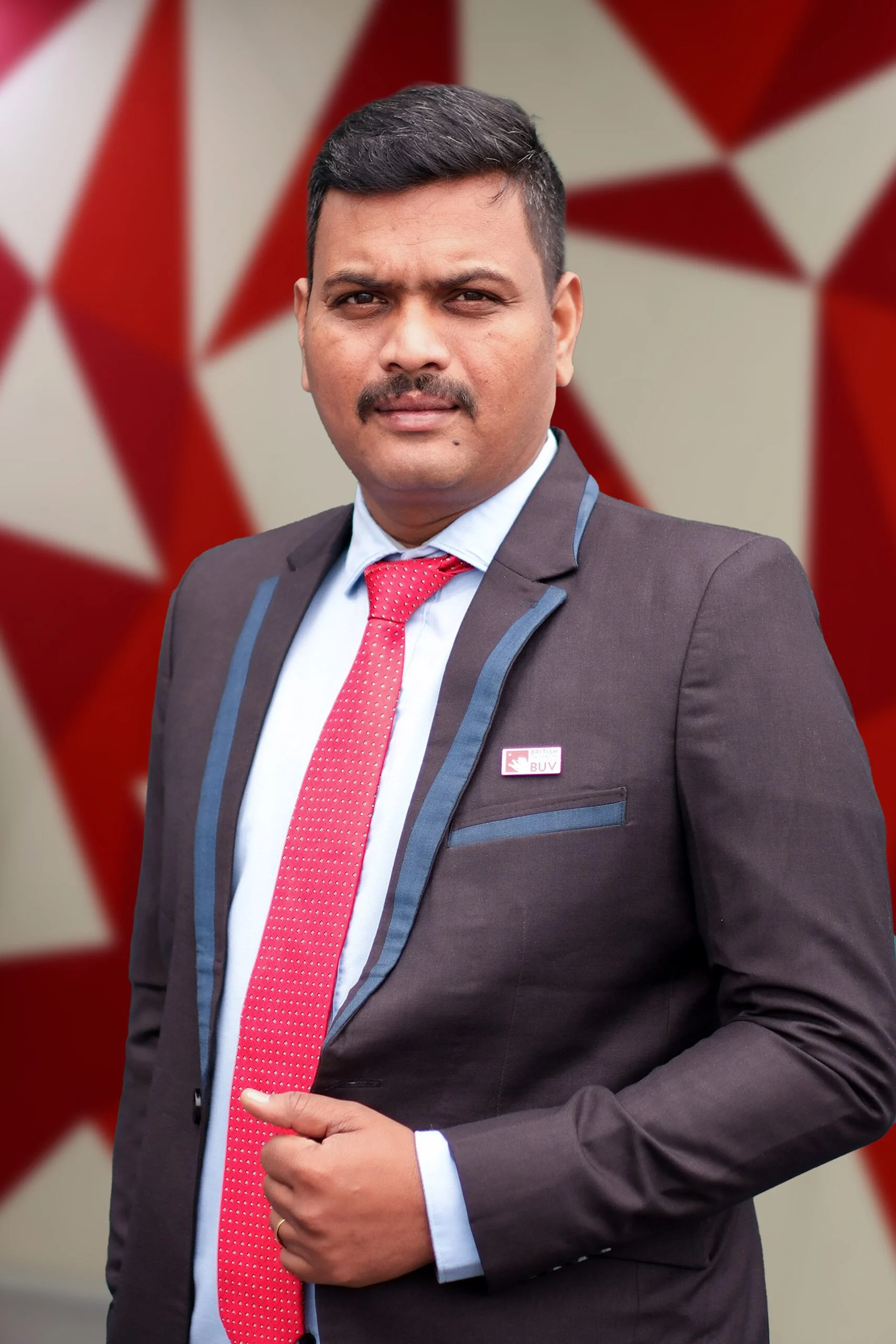 Portrait of a man wearing a dark suit and red tie with a BUV lapel pin, standing against a red-and-white geometric background.
