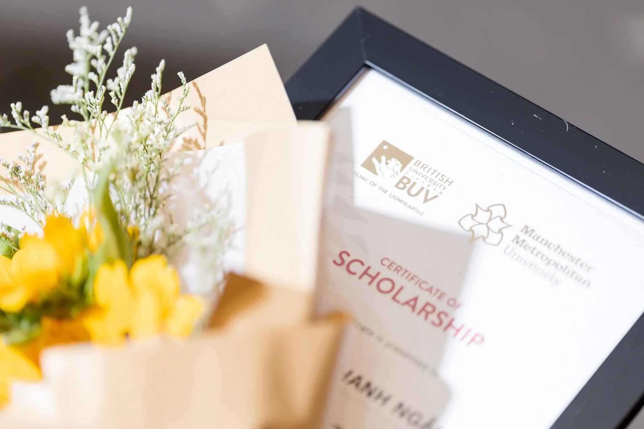 A framed BUV scholarship certificate displayed beside a bouquet of yellow and white flowers.